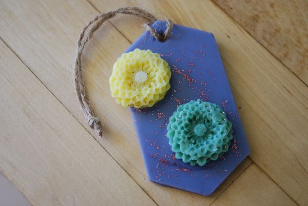 Suspension parfumée ovale de couleur violette avec deux fleurs en cire de couleur verte et jaune agrémentée de paillettes